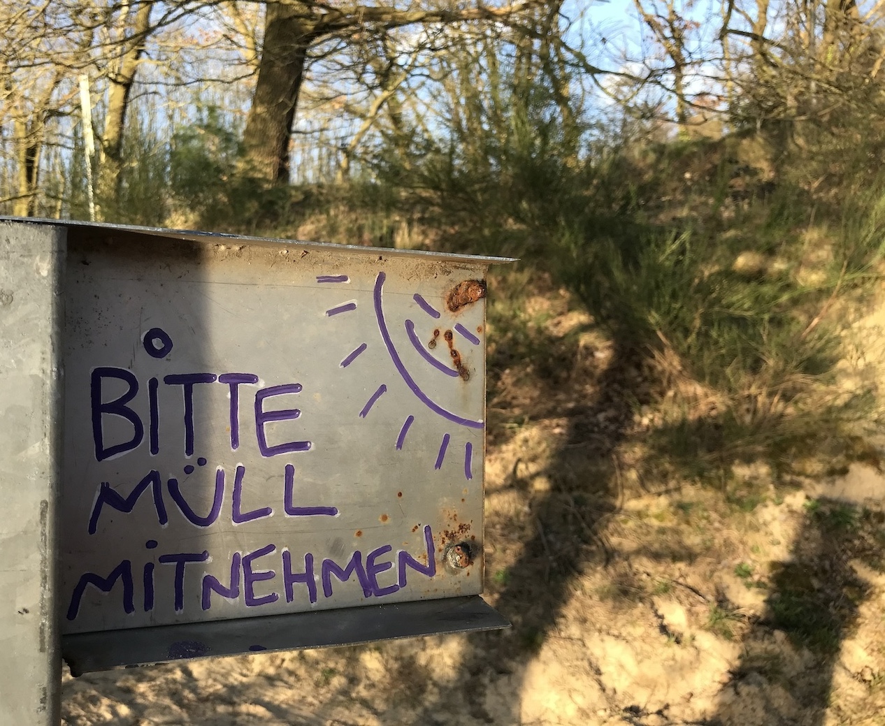 Selbst gemaltes Schild am Strand: „Bitte Müll mitnehmen“. Zu den Worten: freundlich lachende Sonne