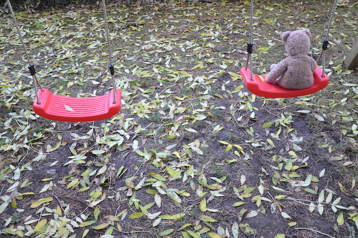 Teddybär von hinten auf einer Schaukel. Daneben: leere Schaukel. Boden ist mit Herbstblättern übersät.