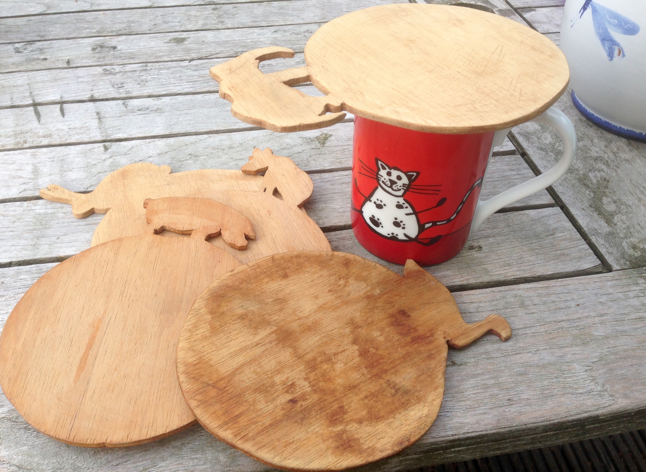 Henkeltasse mit Katzenmotiv, bedeckt mit einer runden, flachen Holzscheibe – Laubsägearbeit. Daneben weitere weitere Laubsäge-Scheiben mit ausgesägten Tiermotiven.