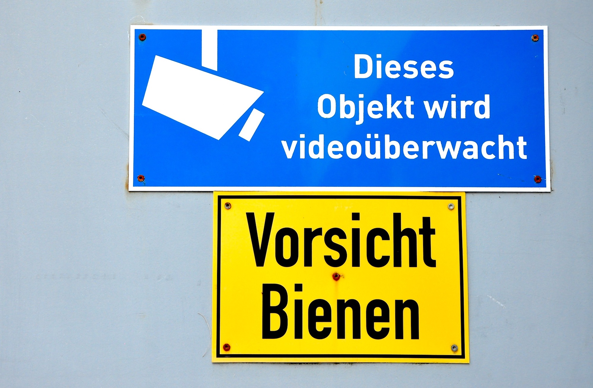 Schild an Stahltür: „Dieses Objekt wird videoüberwacht.“ Darunter gelbes Warnschild: „Vorsicht Bienen“