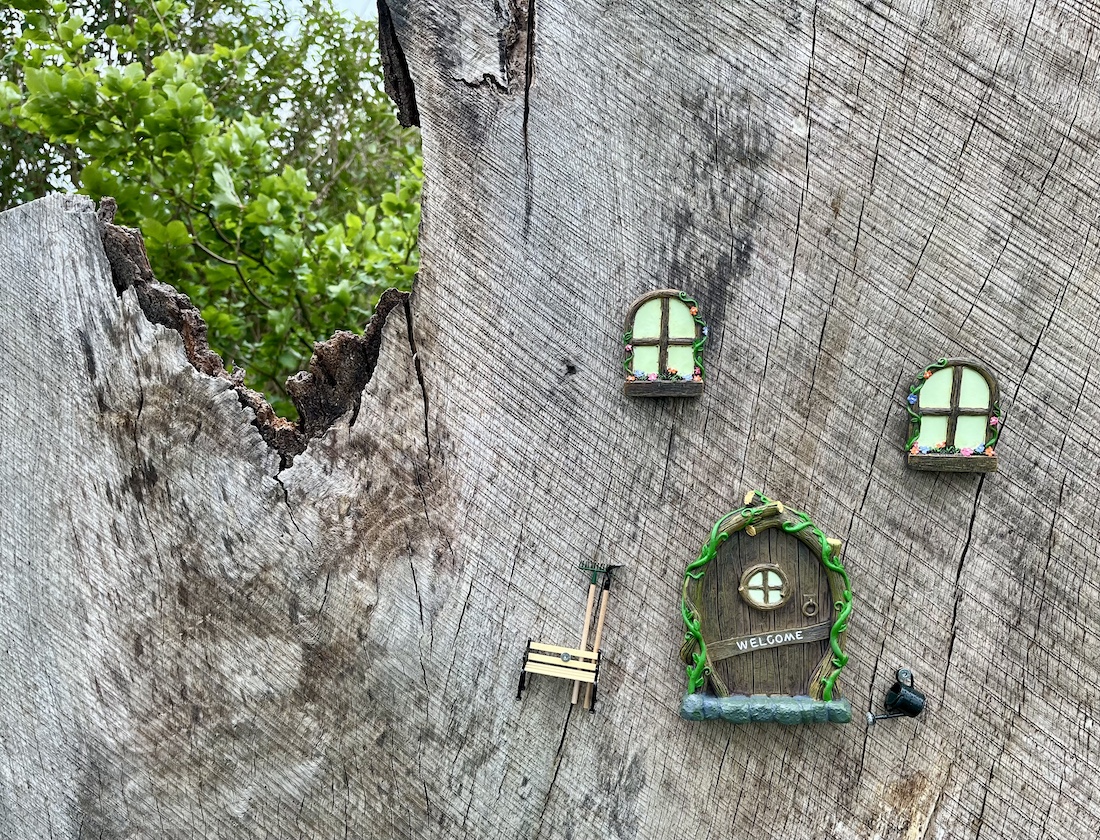 Auf geschliffenem Holzstamm angebrachte kleine Fenster, Tür mit Willkommensschild, Gießkanne und Sitzbank mit Harke